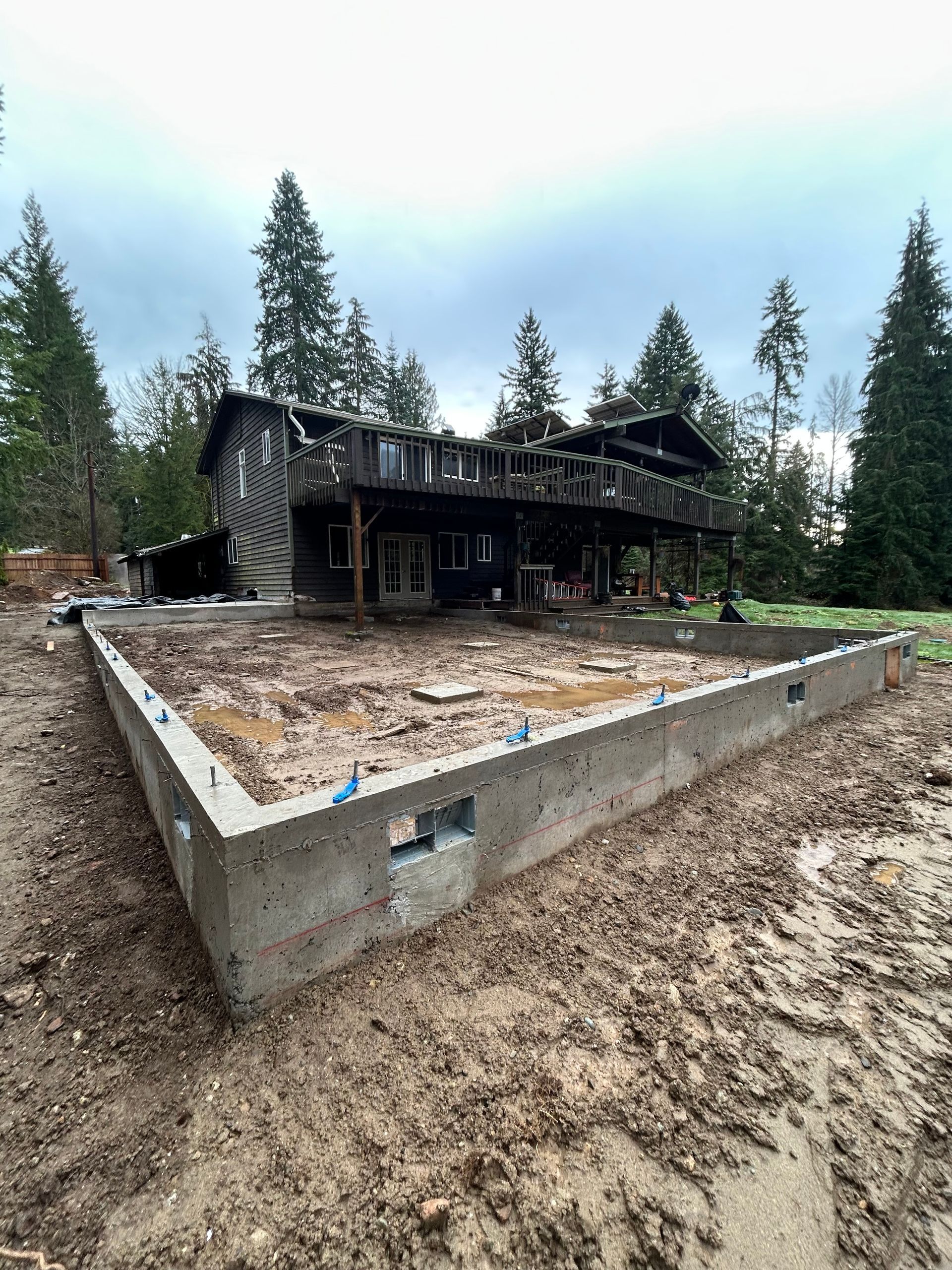 Concrete foundation next to a wooden house with a deck in a muddy yard.