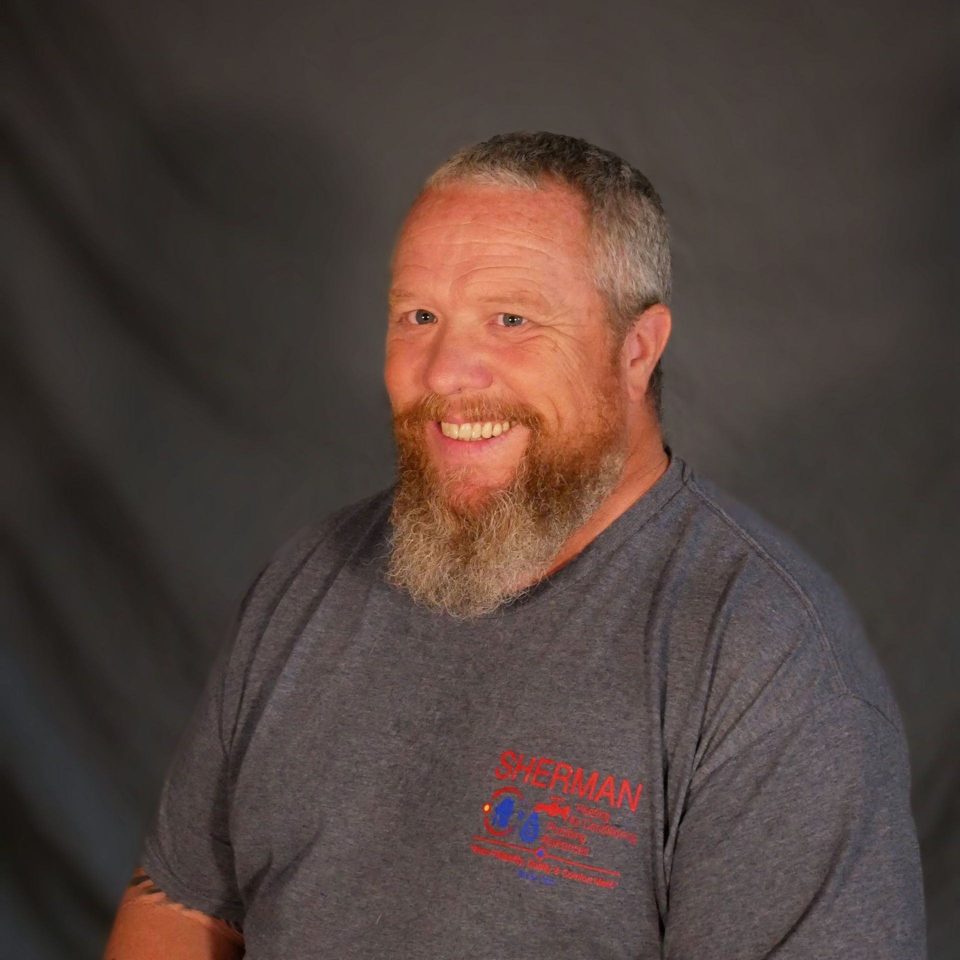 A man with a beard and tattoos is wearing a gray t-shirt and smiling.