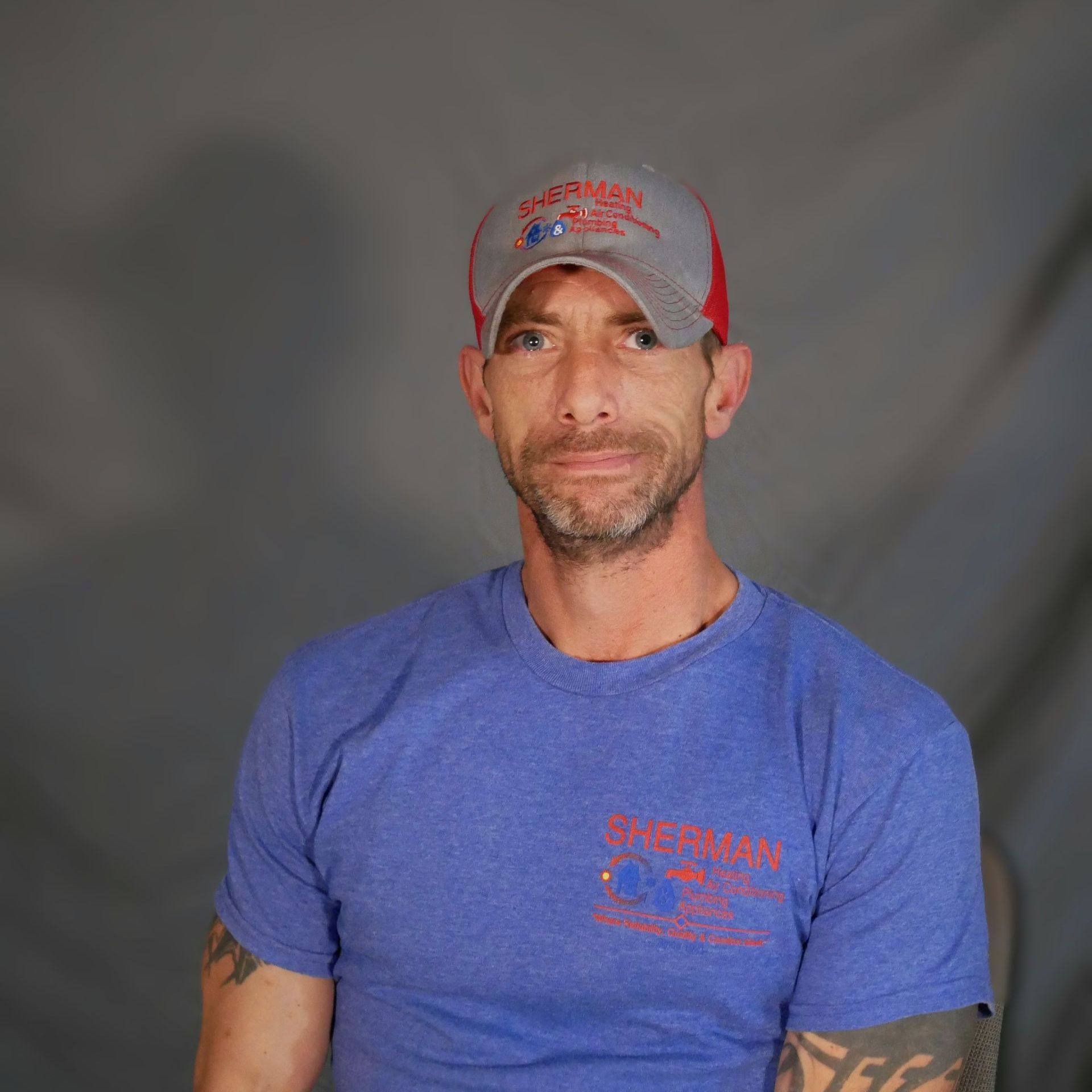 A man with a beard and tattoos is wearing a blue shirt and a hat.