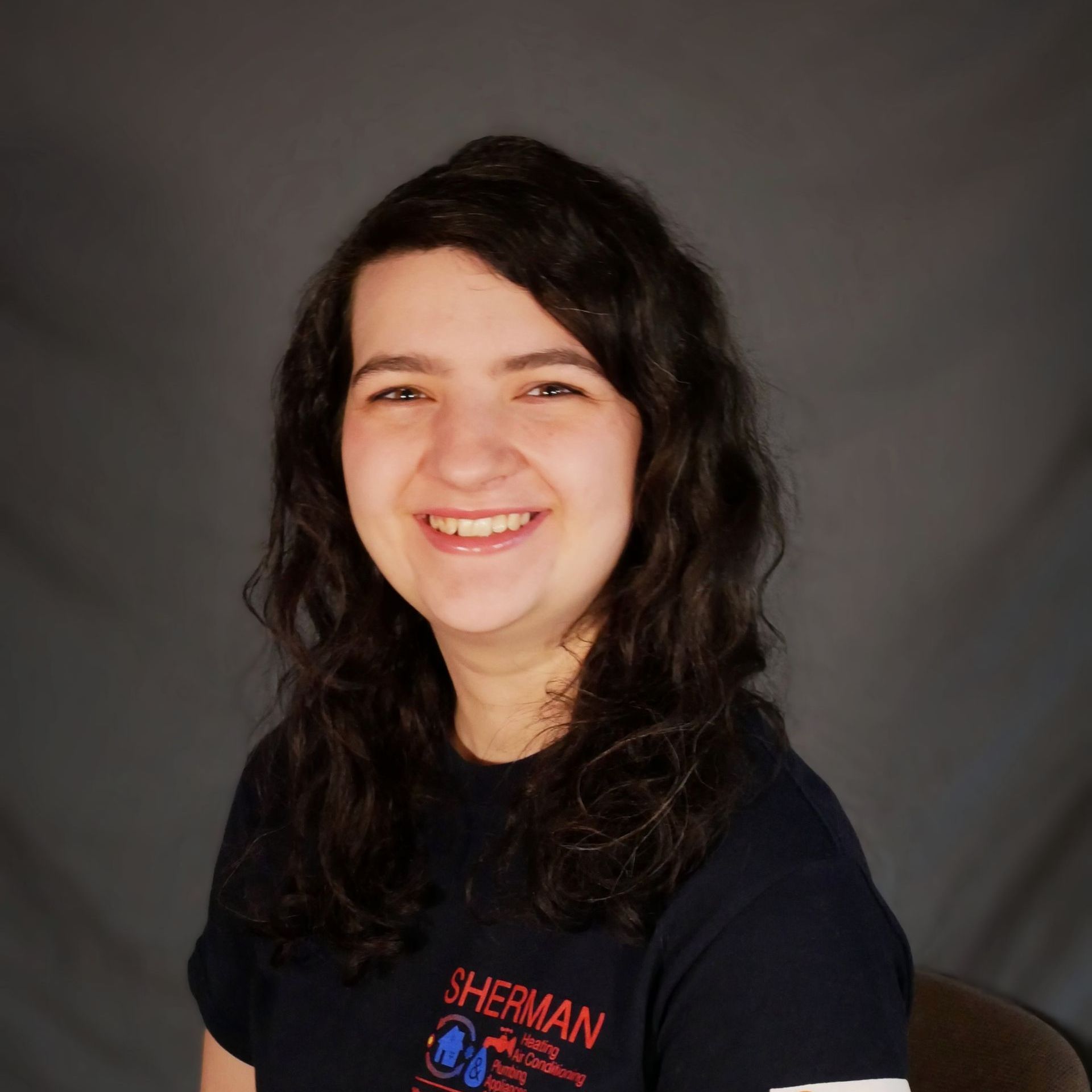 A woman wearing a sherman t-shirt is smiling and sitting in a chair.