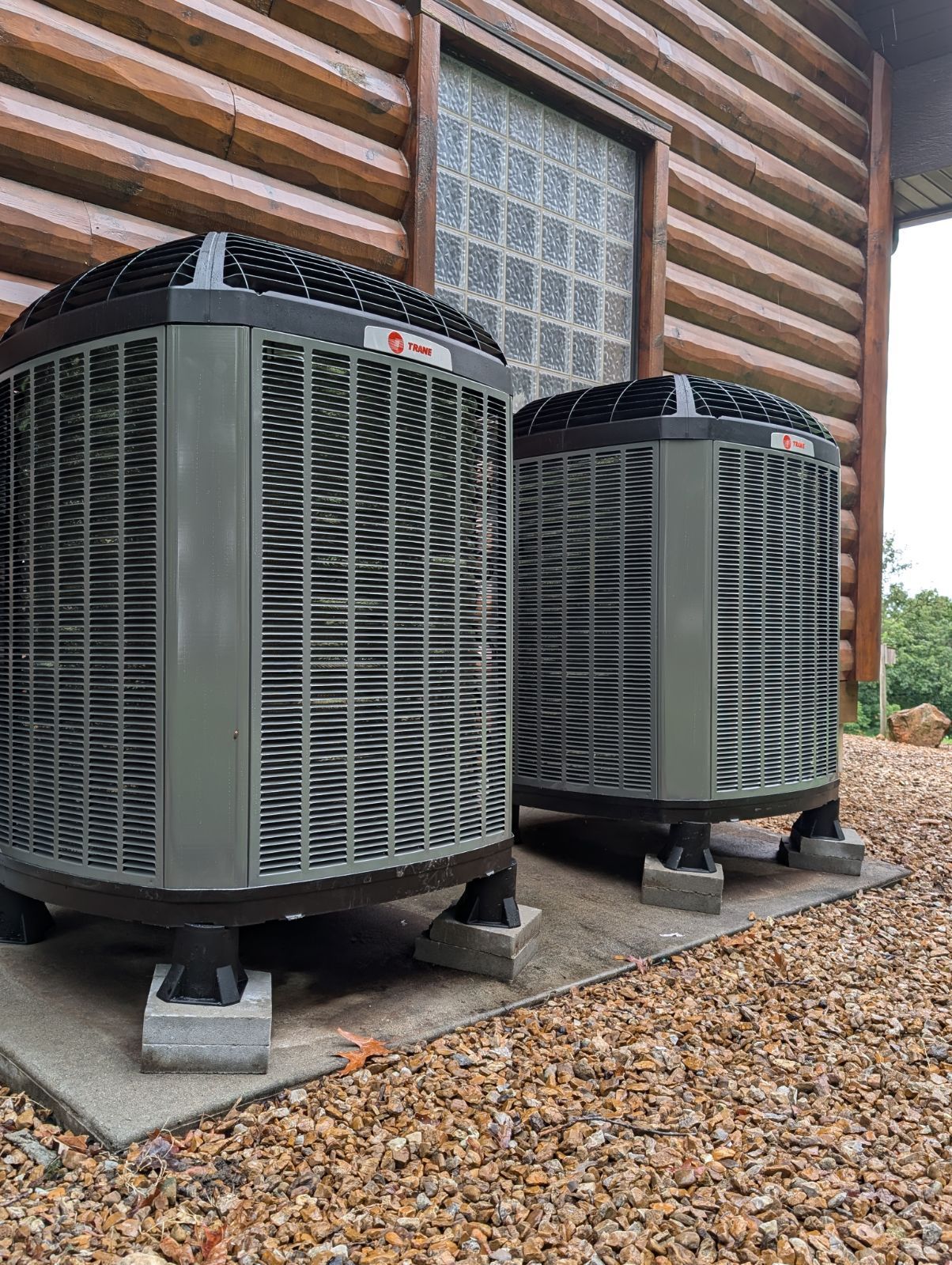 Two gray HVAC units on concrete pads near a log cabin.