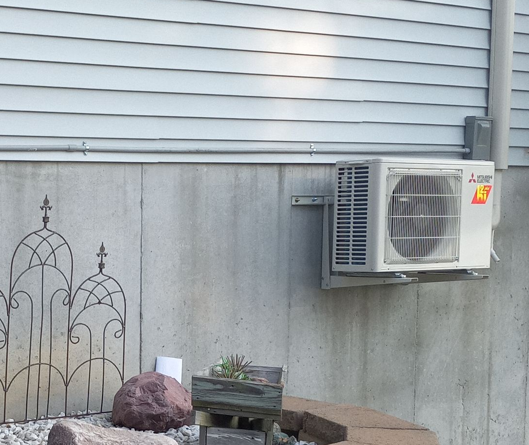 Wall-mounted Mitsubishi air conditioning unit on a concrete wall. Siding above, metal art and landscaping below.