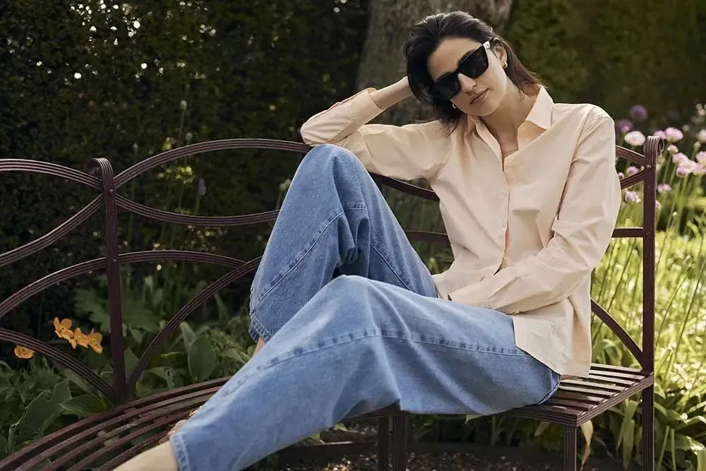 En kvinde sidder på en bænk iført solbriller og jeans.