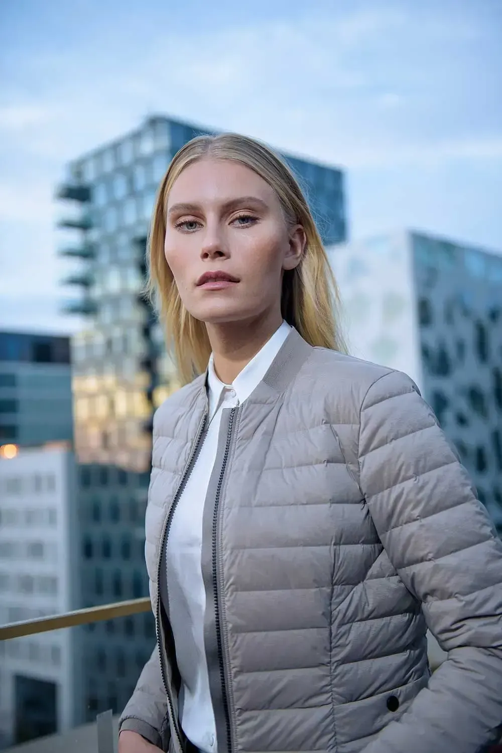 En kvinde iført bomberjakke og hvid skjorte står foran en bys skyline.