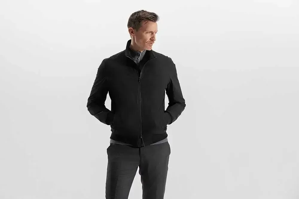 En mand iført sort jakke og grå bukser står med hænderne i lommerne.