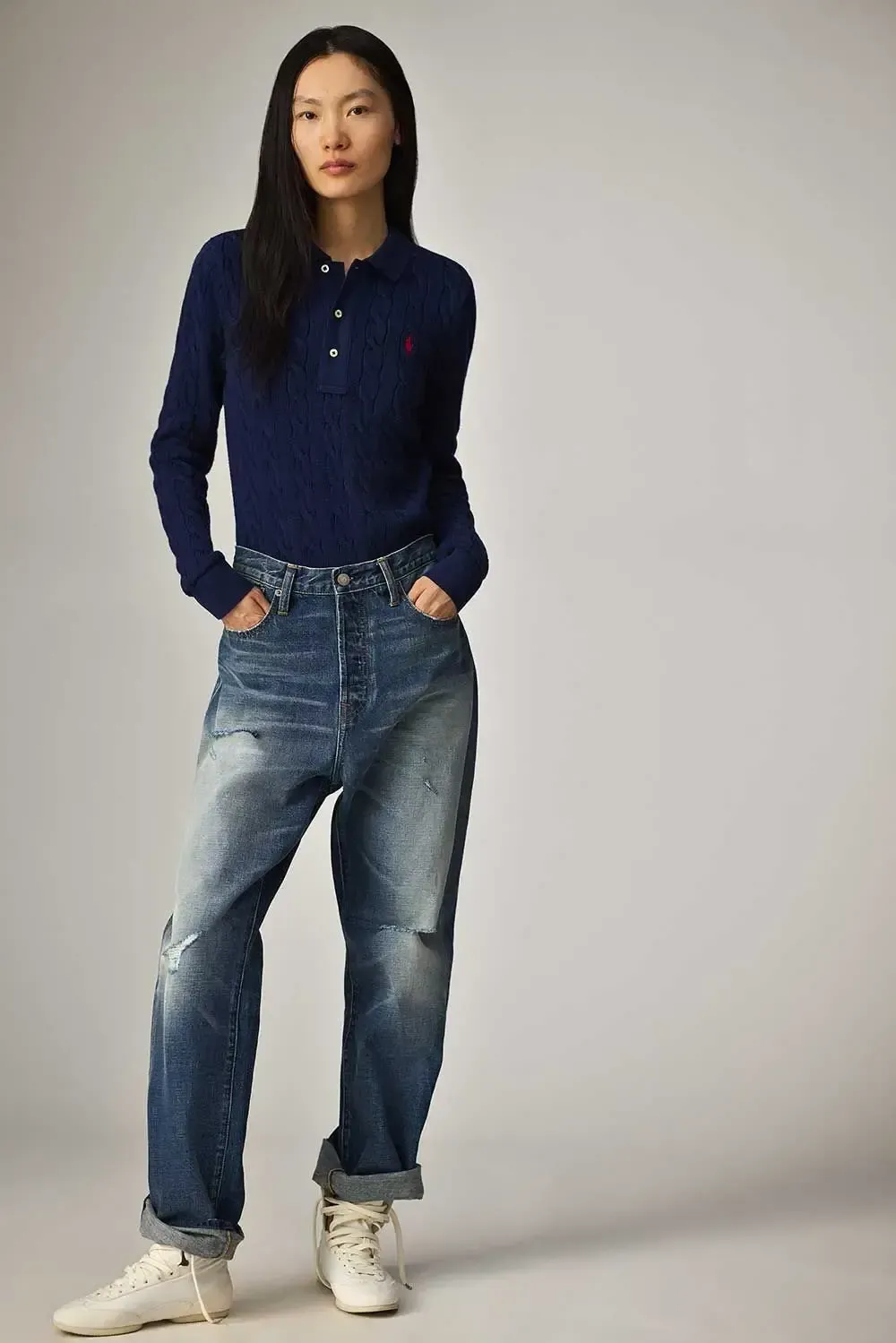 En kvinde iført en blå poloshirt og jeans står med hænderne i lommerne.