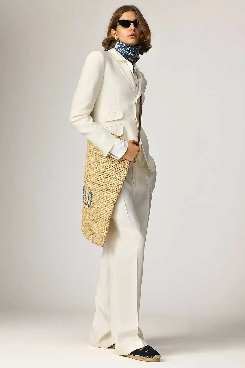 En model iført et hvidt jakkesæt og solbriller holder en stråtaske.