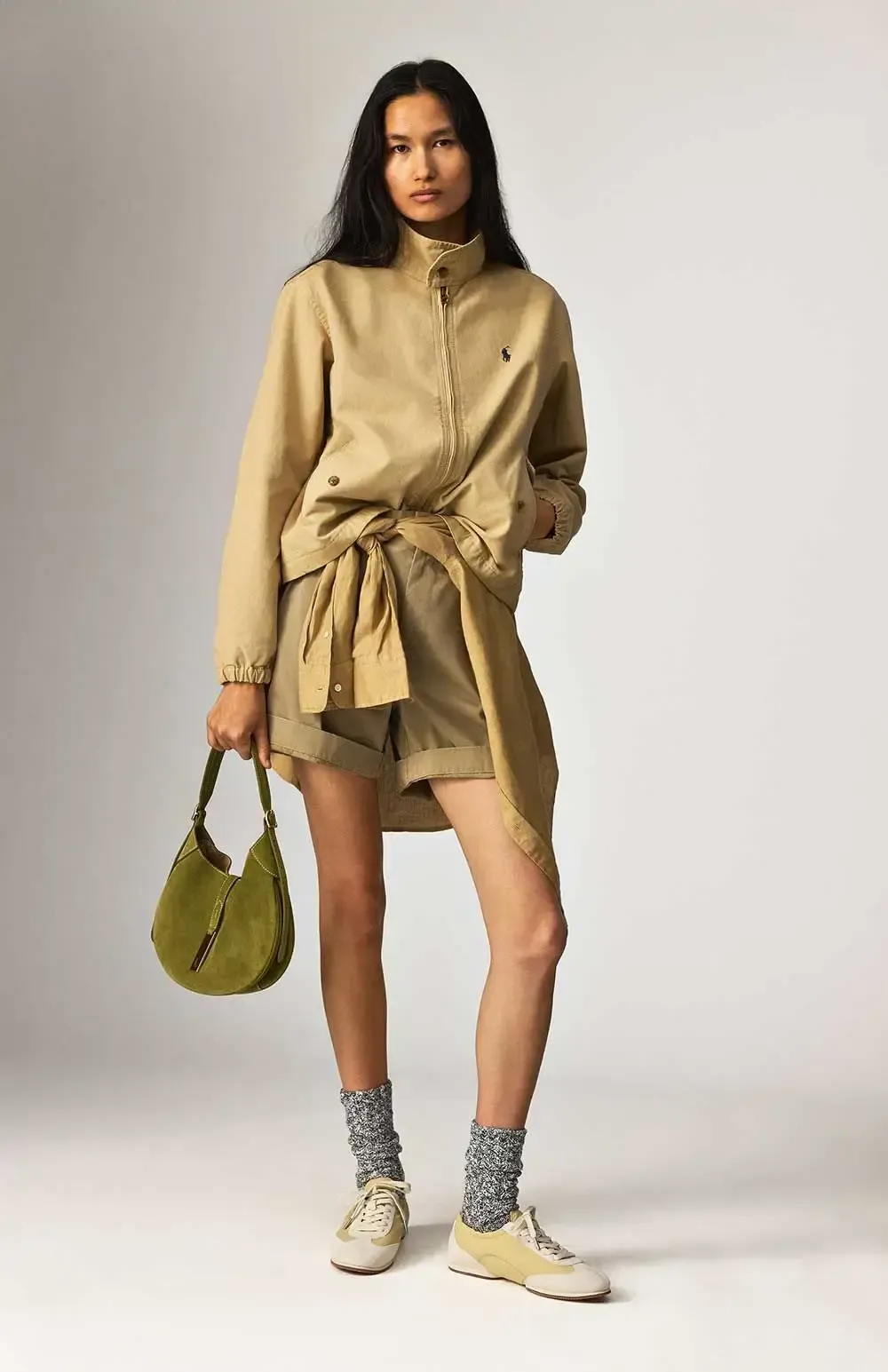 En kvinde er iført en brun jakke og shorts og holder en grøn taske.