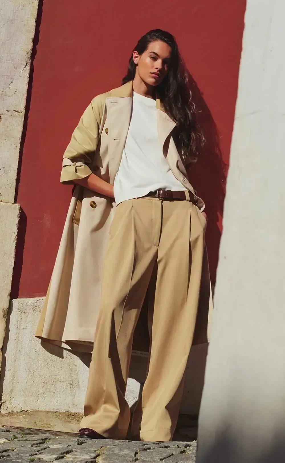 En kvinde iført trenchcoat og bukser med vide ben står foran en rød væg.