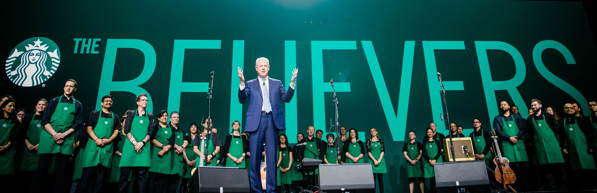 A man stands on a stage in front of a sign that says the believers