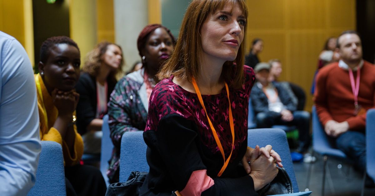 A focused view of a woman listening attentively in an audience. She sits upright with a slight smile on her face.