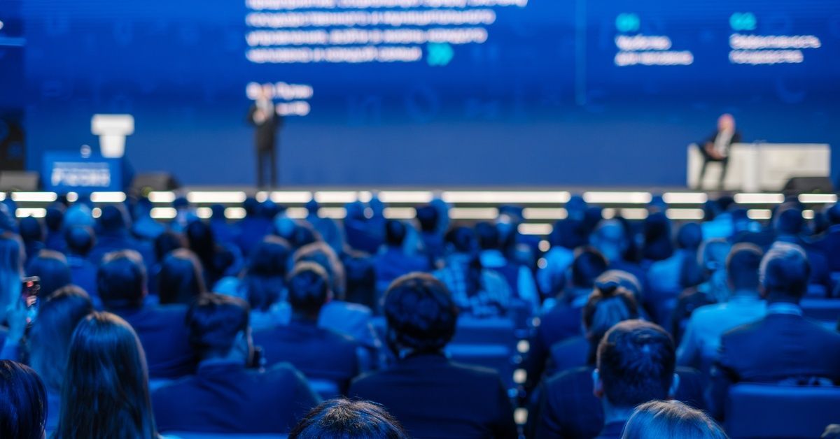 View from behind an audience watching a presenter at a professional event. Blue light illuminates th