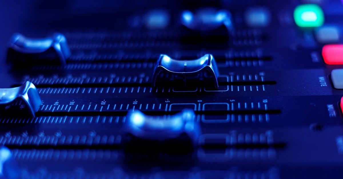 Close-up view of vertical sliding faders on a sound mixing console. Green and red lights are lit up on the board.