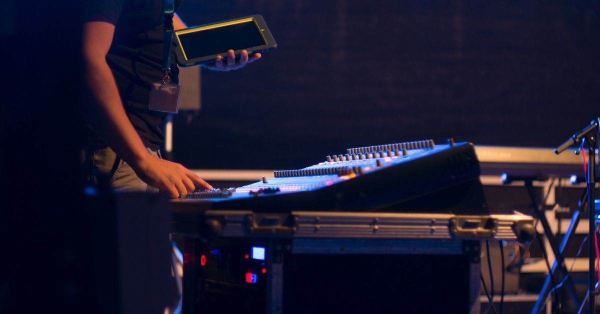 A technician adjusts controls on a large audio mixing console in a dimly lit space. They hold a tablet in one hand.