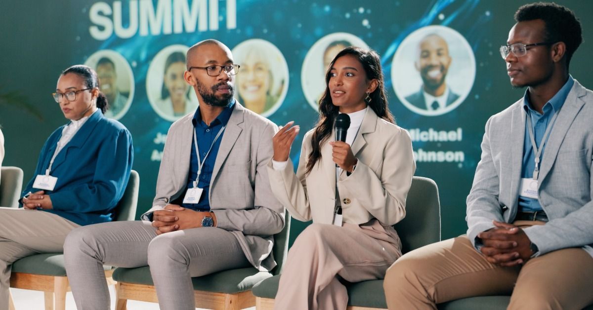Four panelists sit on stage at a conference summit in front of a branded backdrop. One speaks into a wireless microphone.