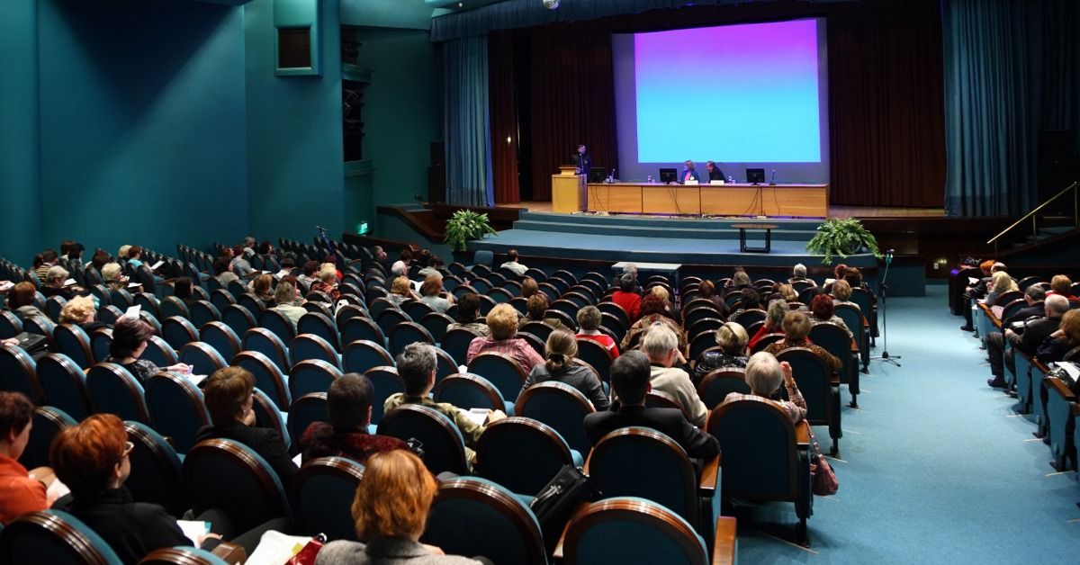 On a stage, one person stands at a podium and two sit at a table in front of a projection screen.