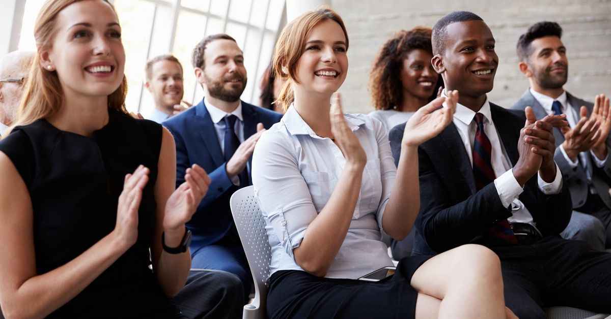 An audience dressed in business attire smiles and applauds. Behind them, floor-to-ceiling windows let in natural light.