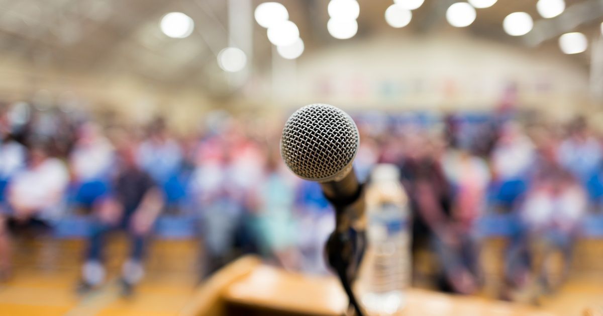 A close-up of a microphone positioned at a podium, with a blurred audience seated in the background 