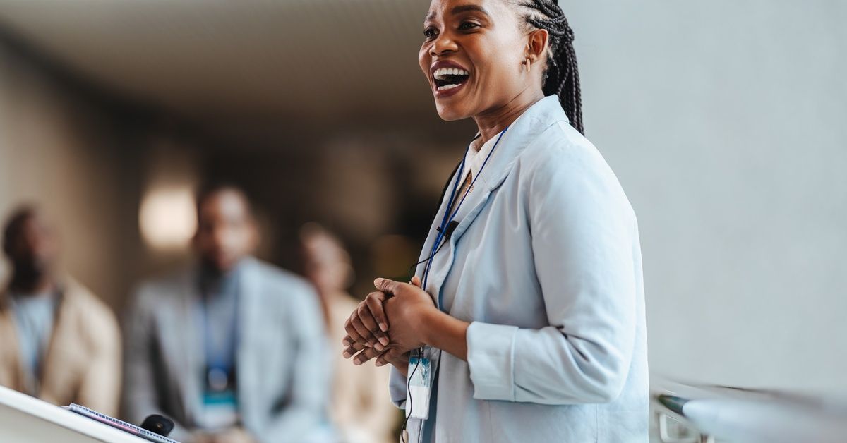 Side view of a woman in a blazer speaking with a microphone clipped to her lapel. The event attendees are blurred.