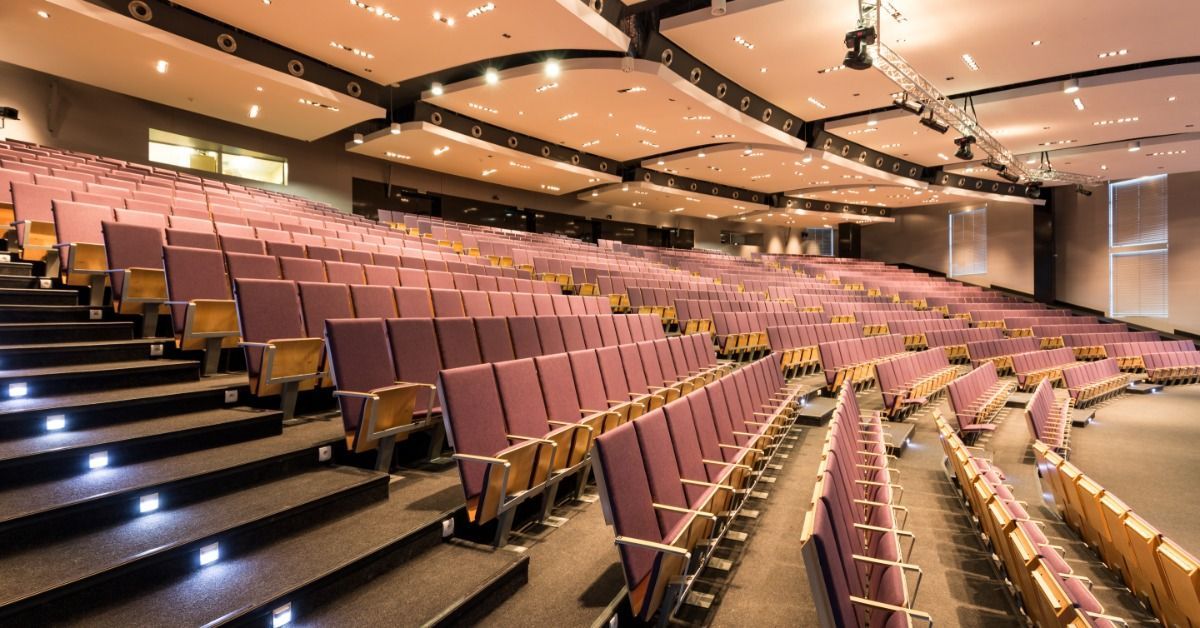 An empty venue has tiered seating, carpeted aisles, and upholstered chairs. Large ceiling panels hold recessed lights.