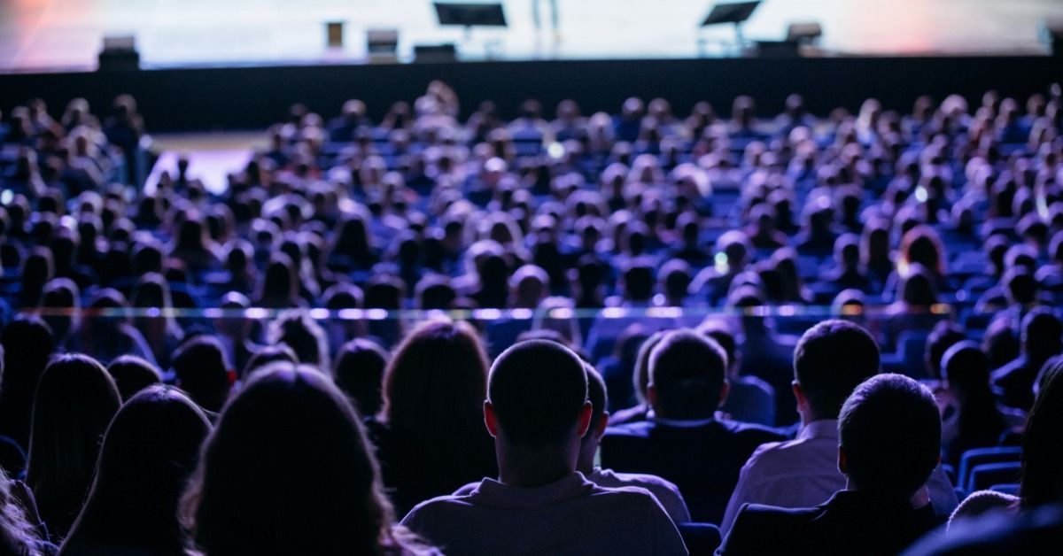 A large audience in the foreground faces a brightly lit auditorium stage. The speaker appears small in the distance.