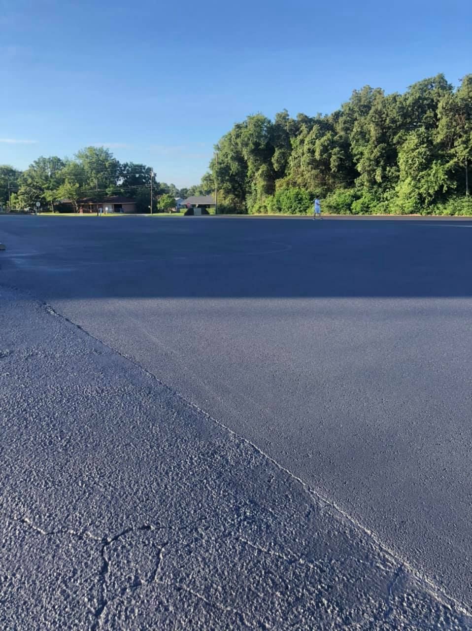 A freshly paved, dark asphalt parking lot under a bright blue sky, with trees in the background.