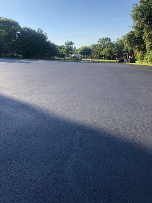 Newly paved, dark asphalt parking lot on a sunny day, surrounded by green trees.