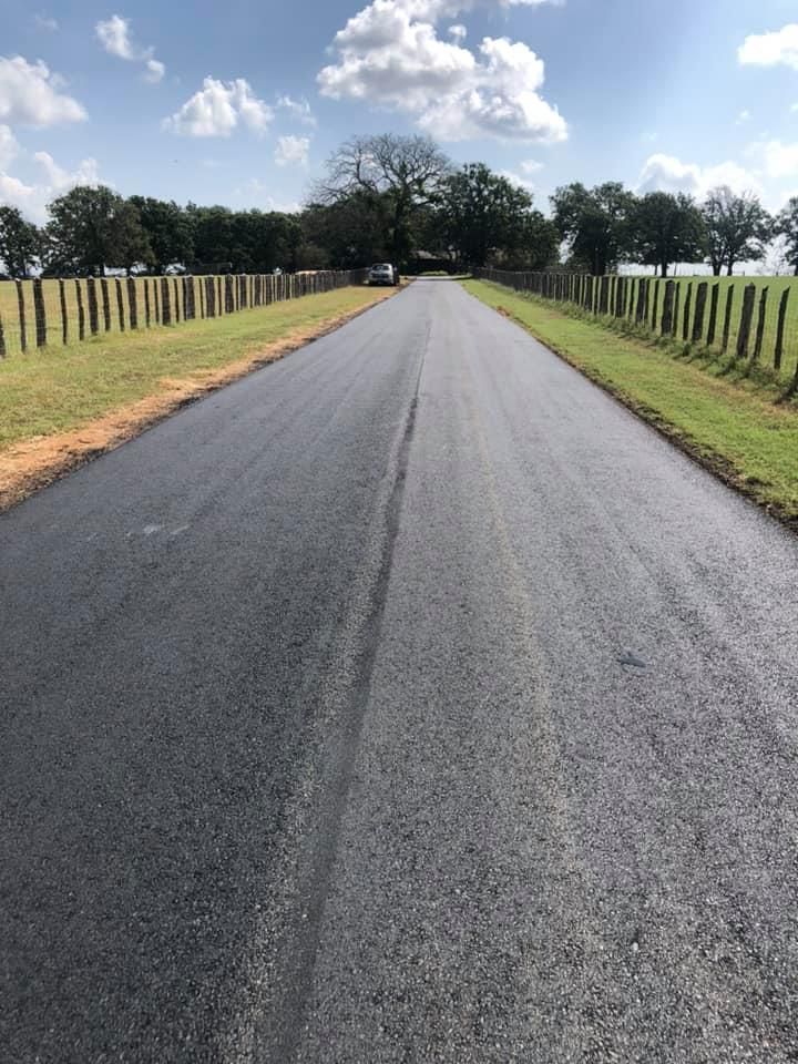 Paved road leading through a rural landscape with a fence on either side, blue sky with clouds in the background.