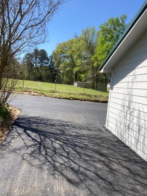 Asphalt driveway next to a white building, tree shadows, green lawn, and trees in the background.