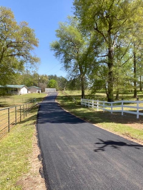 Asphalt driveway leading to a white fence and houses under a blue sky.