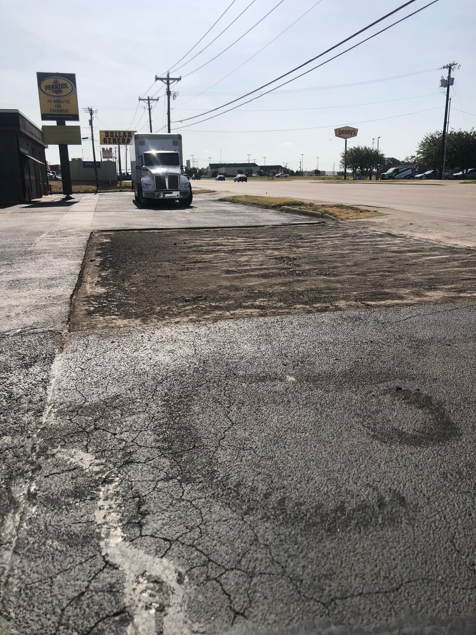 Damaged asphalt with a semi-truck parked, power lines above, and businesses in the distance.