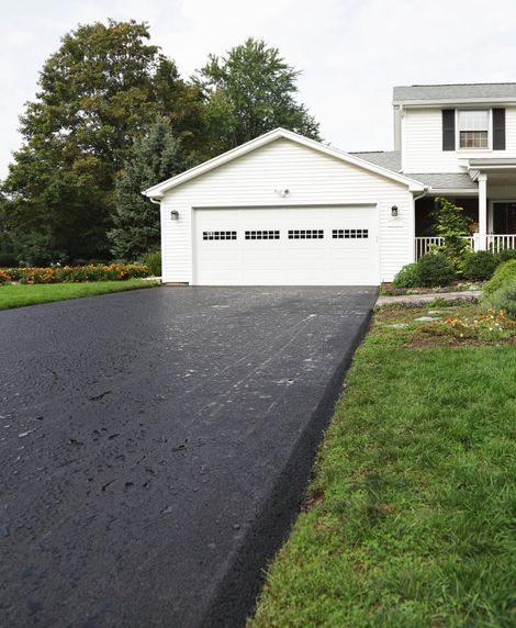 Black asphalt driveway leading to a white garage of a house, bordered by green grass and landscaping.