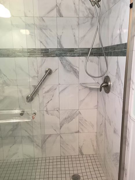 Shower stall with marble-patterned tile, glass door, grab bar, showerhead, and small mosaic accent.