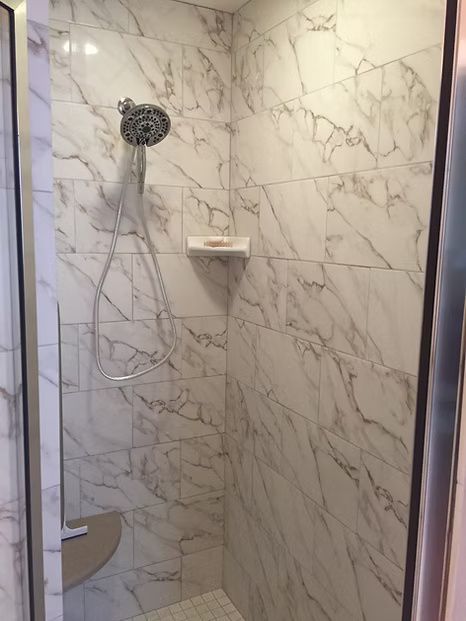 Shower with white marble-patterned tile, handheld showerhead, and corner shelf.