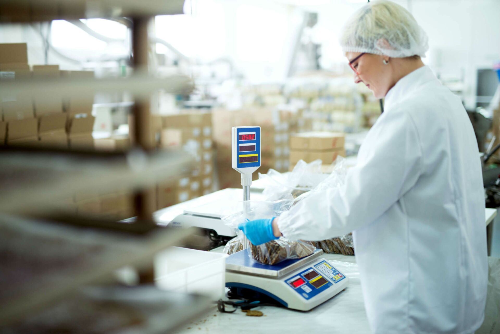 Une personne vêtue d'une blouse blanche et d'une charlotte utilise une balance numérique pour peser un produit dans une usine de fabrication.