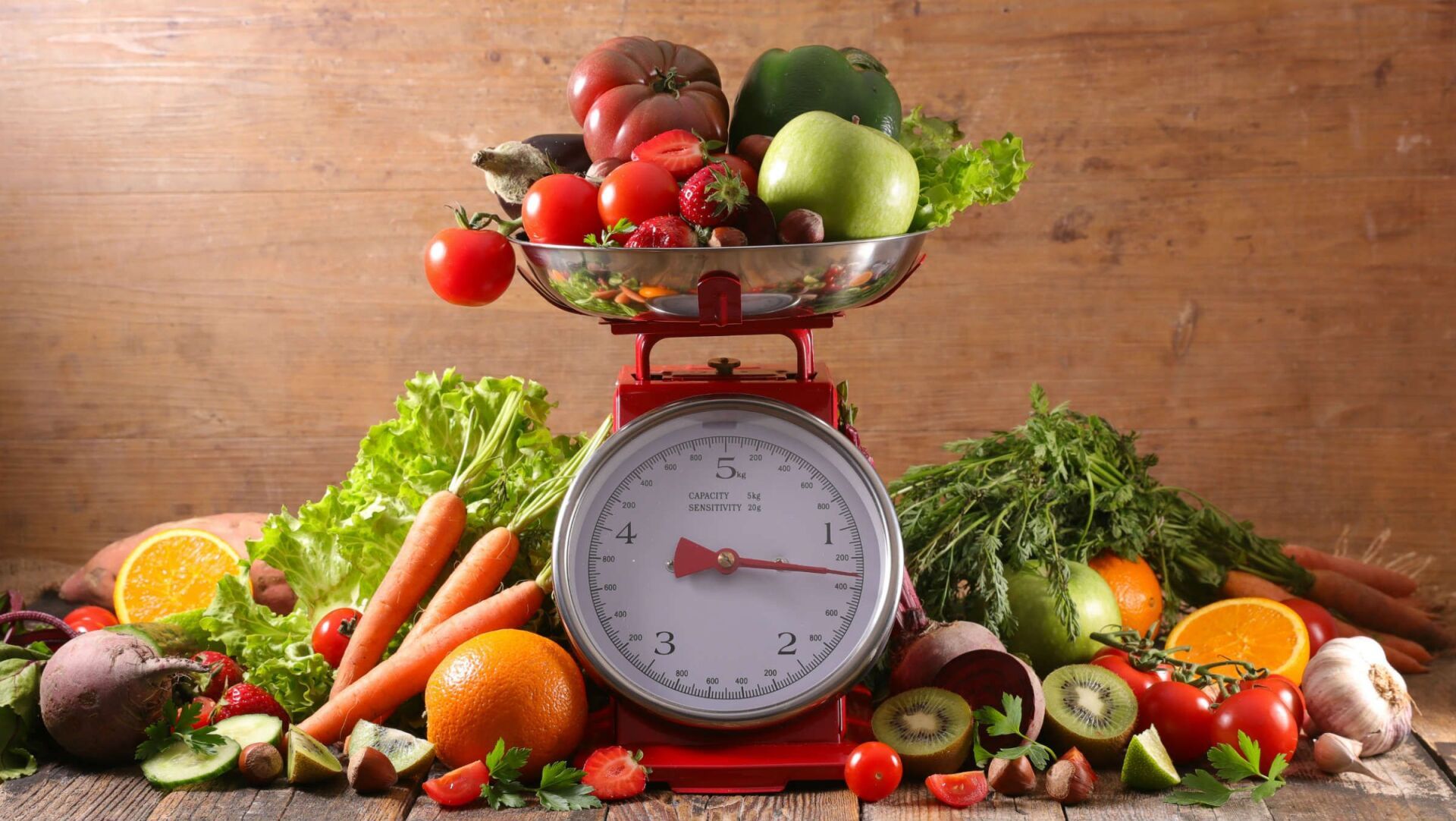 Une balance de cuisine rouge est posée sur une surface en bois, recouverte d'une épaisse pile de fruits et légumes frais comme des pommes, des baies et des carottes.