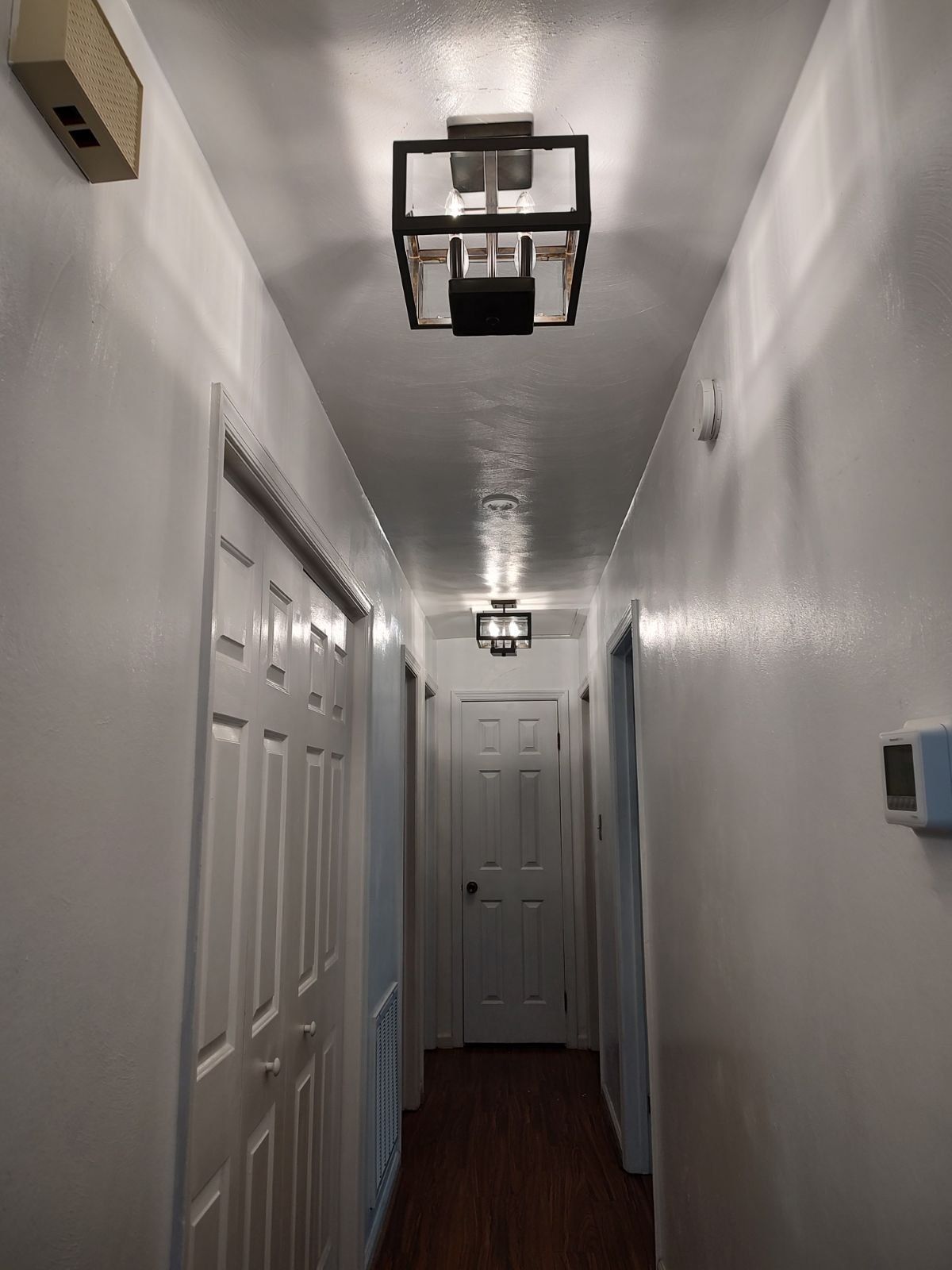 Narrow hallway with white walls and ceiling, dark wood floor, and three square light fixtures.