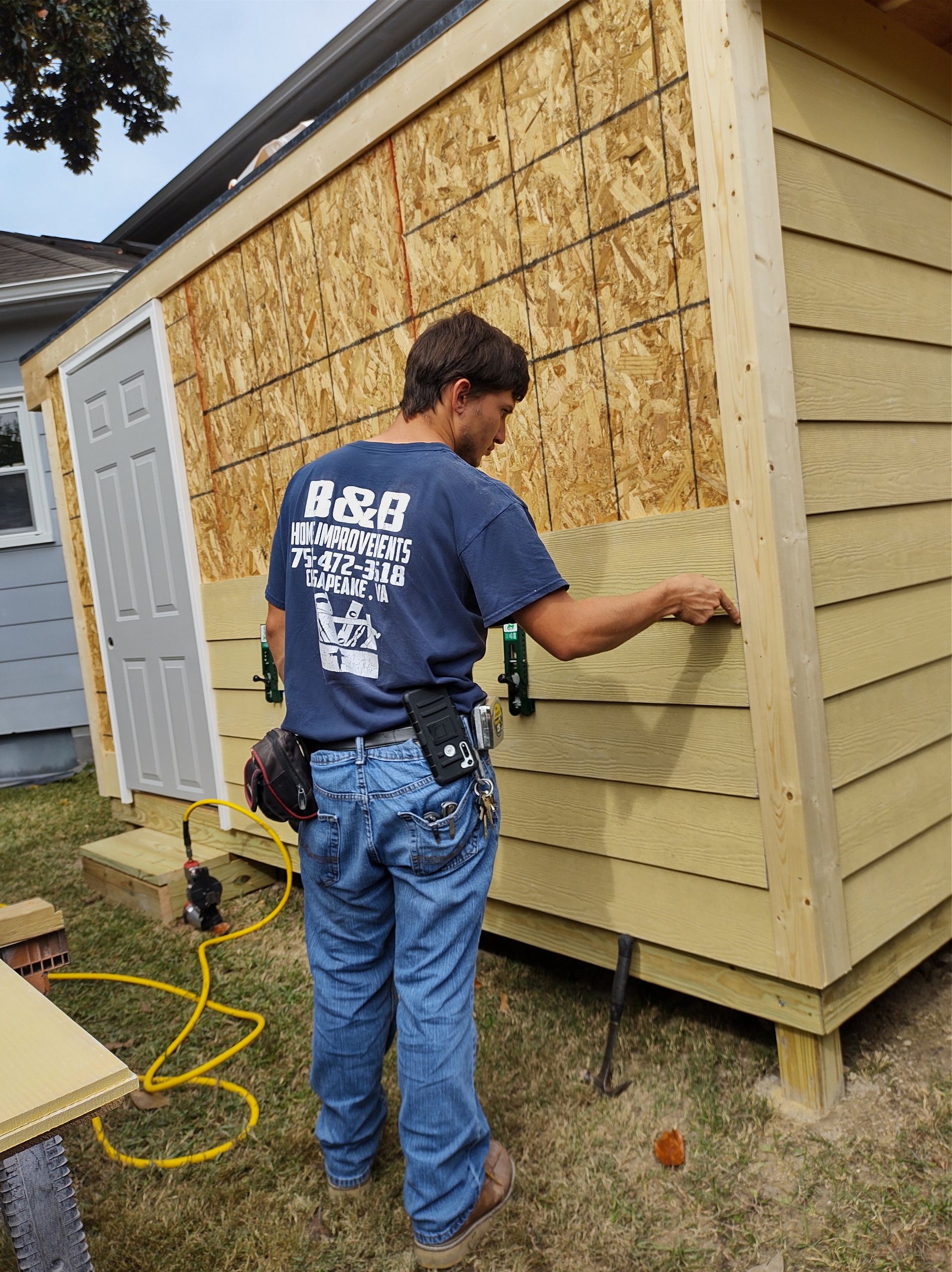 A construction worker in blue shirt and jeans installs siding on a shed in a yard.