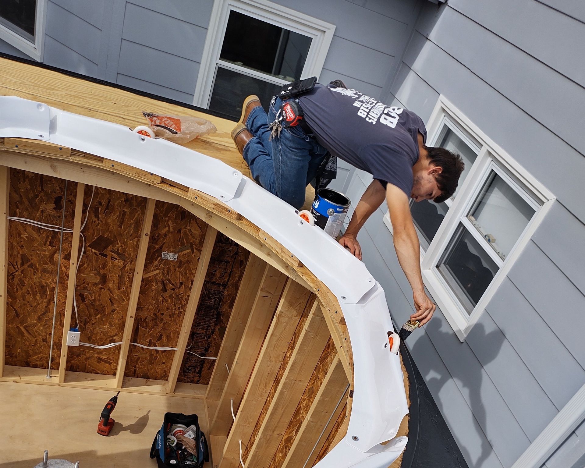 Two roofers working on a curved roof section of a house; gray siding and white windows.