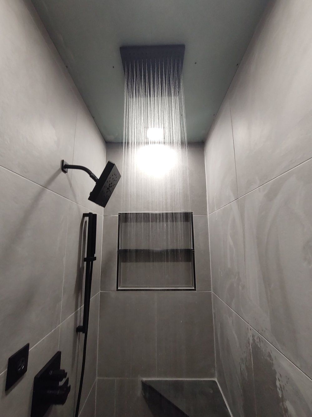 Black showerhead and rain showerhead in a gray tiled shower, water pouring.
