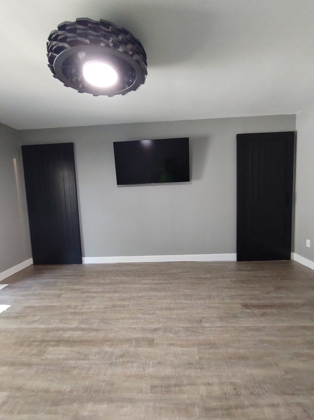 A room with gray walls, wood flooring, a mounted TV, black doors, and a decorative ceiling light.