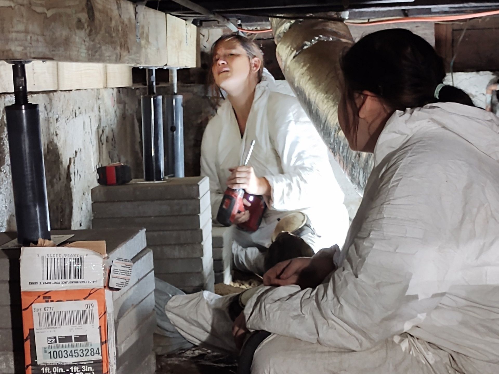 Two people in white coveralls work under a house. One holds a tool, inspecting support beams.
