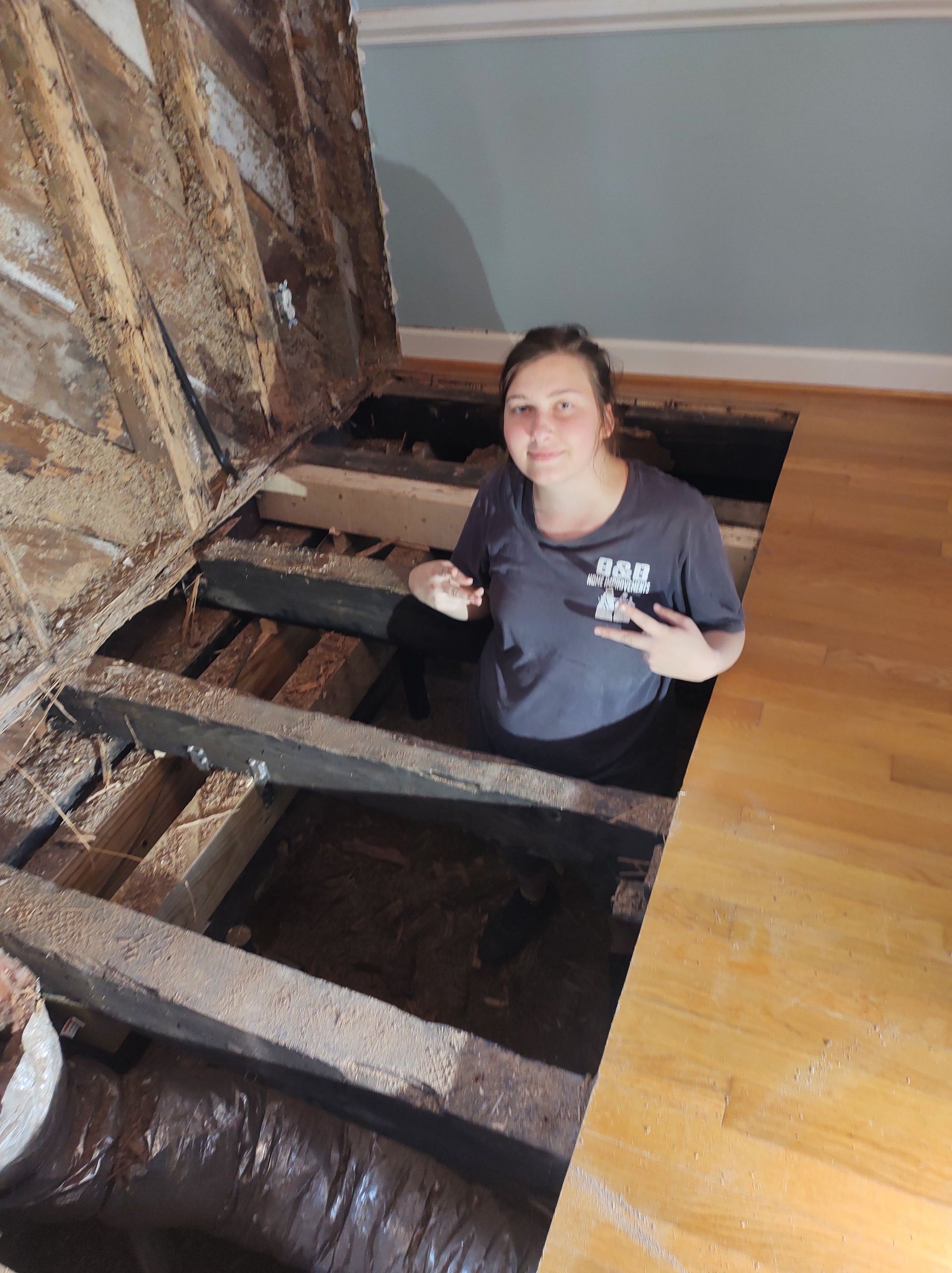 Woman in open floor, giving peace sign. Construction debris, wood beams, light wood floor, blue walls.