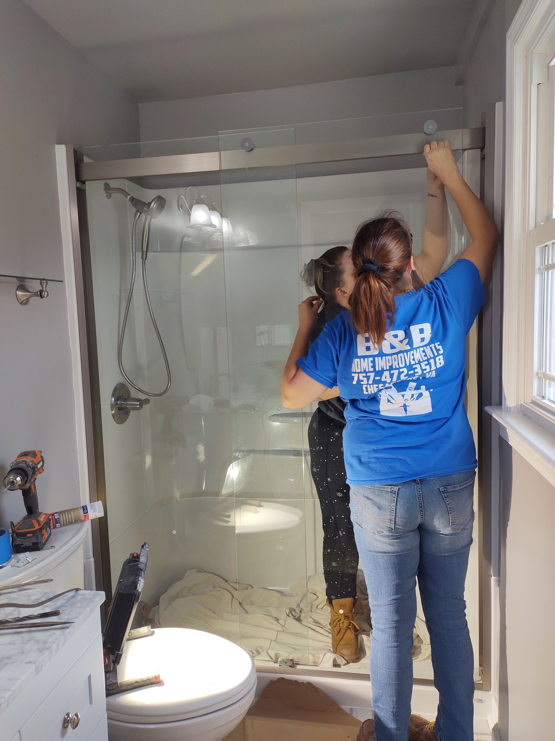 Two people installing a shower door in a bathroom. One in a blue shirt is reaching up.