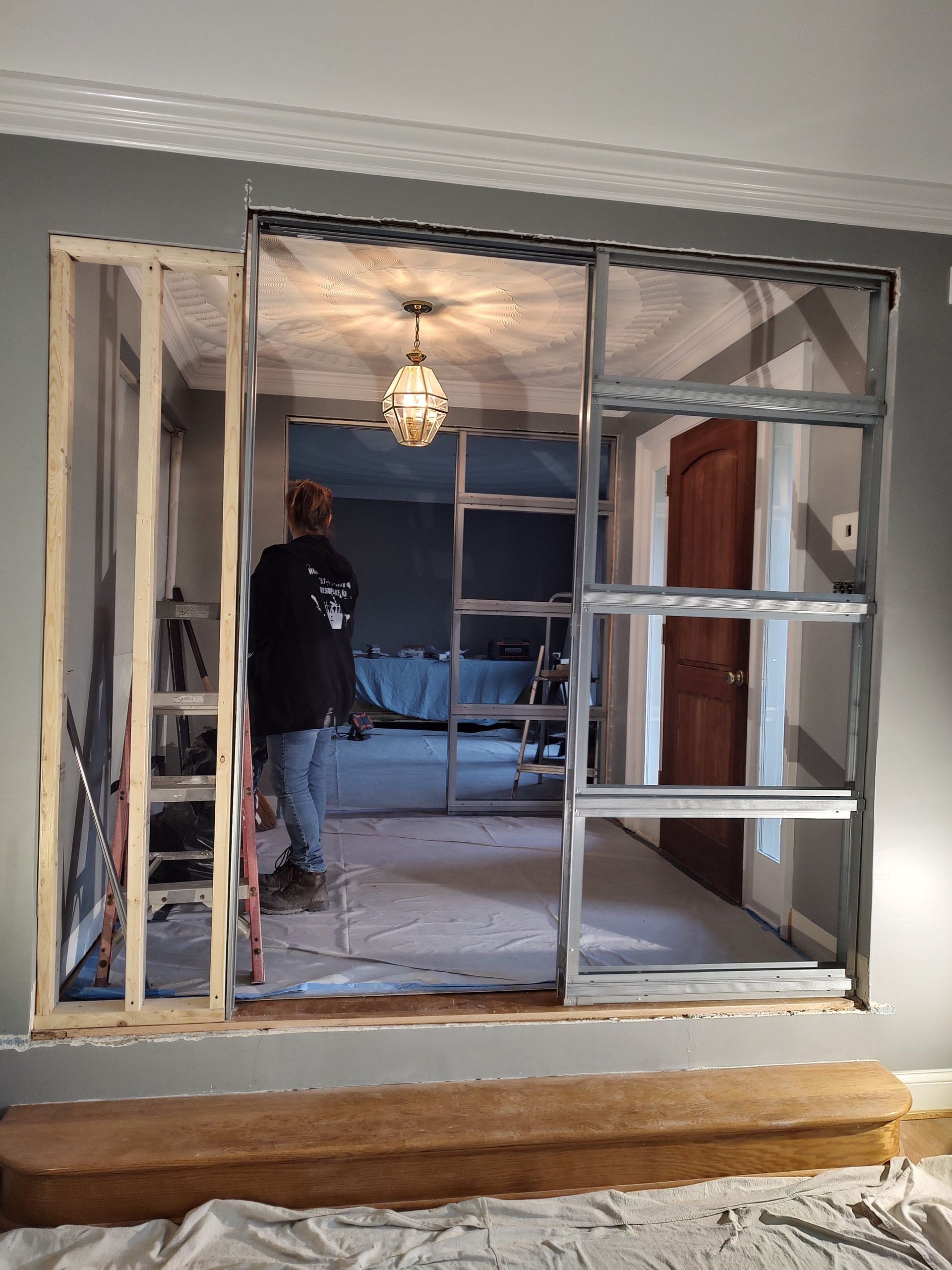 Person framed by interior wall during renovation; metal studs, gray walls, brown door visible.