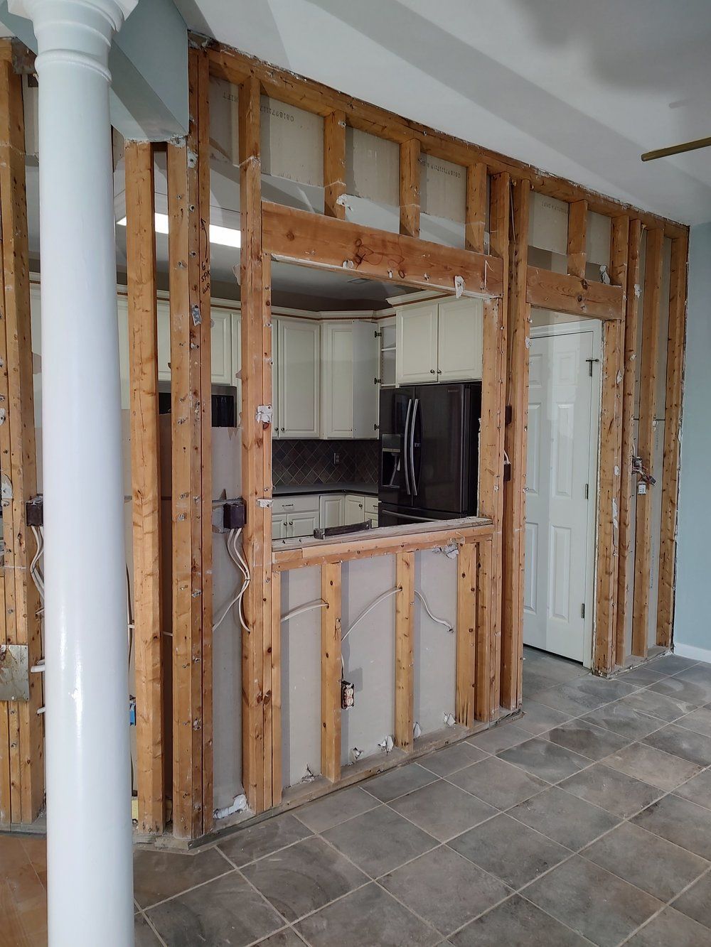 Interior wall under construction; open view to kitchen. Studs visible. White cabinets, black fridge.
