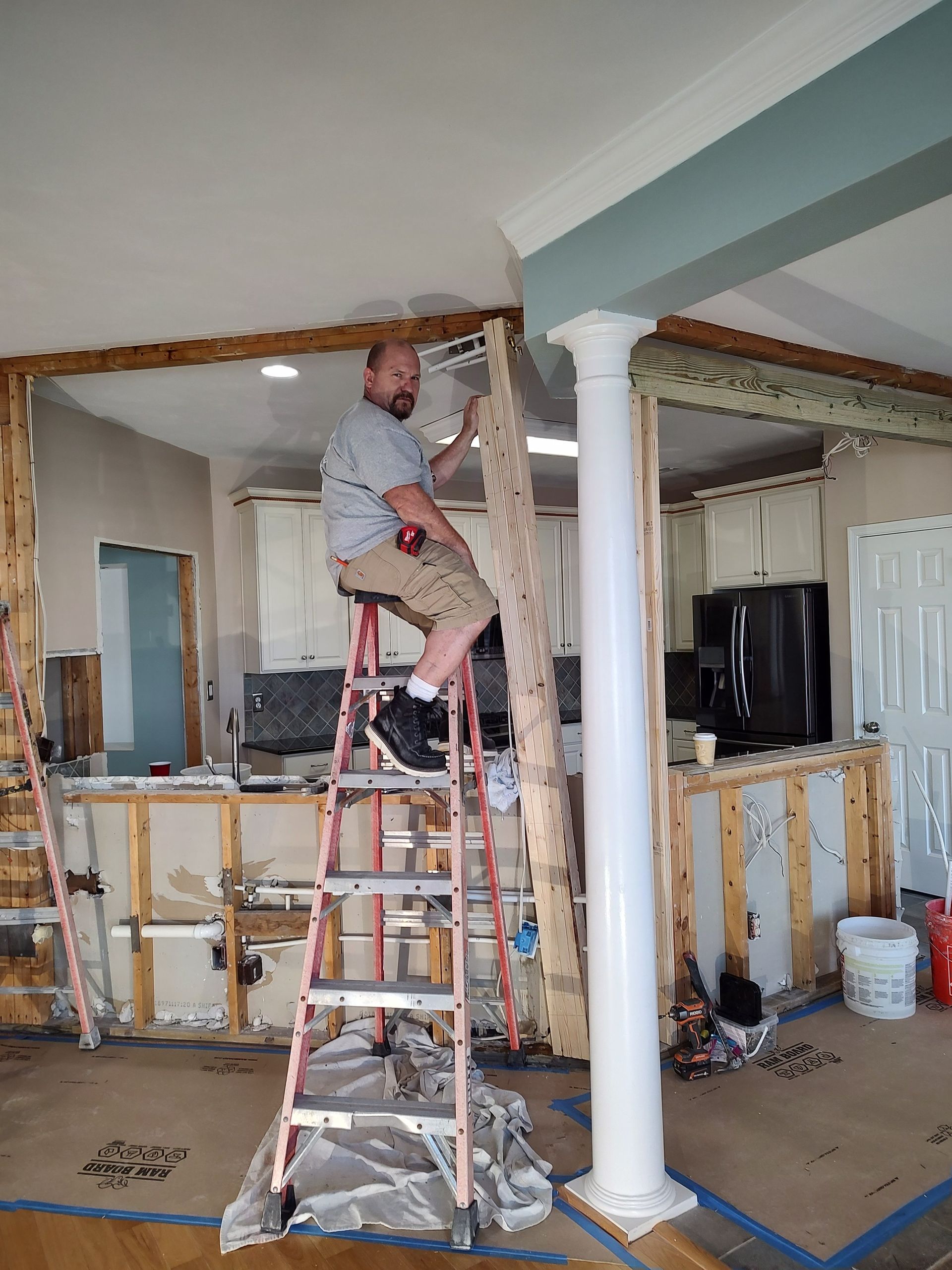 Man on a ladder removes wood trim in a kitchen renovation. The space has exposed studs and a white column.