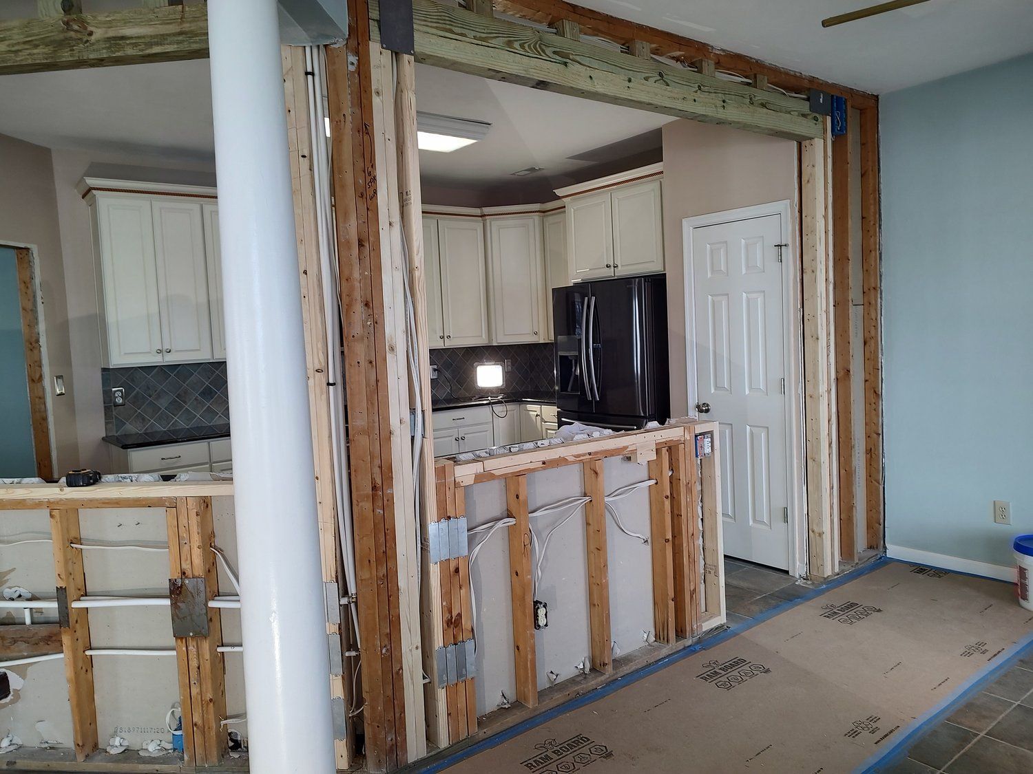 Interior renovation showing a partial wall removed between a kitchen and living area; exposed studs, electrical wires, and cabinets.