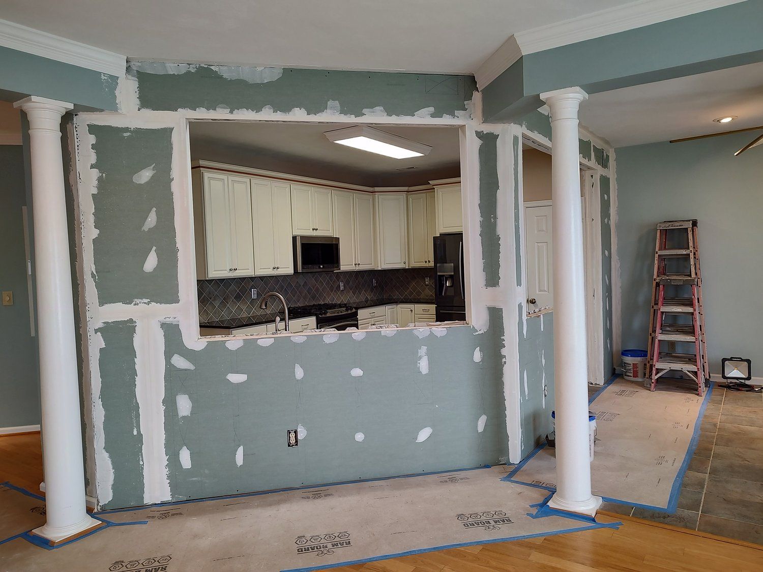 Room under renovation with a partially opened wall, kitchen visible beyond the opening.
