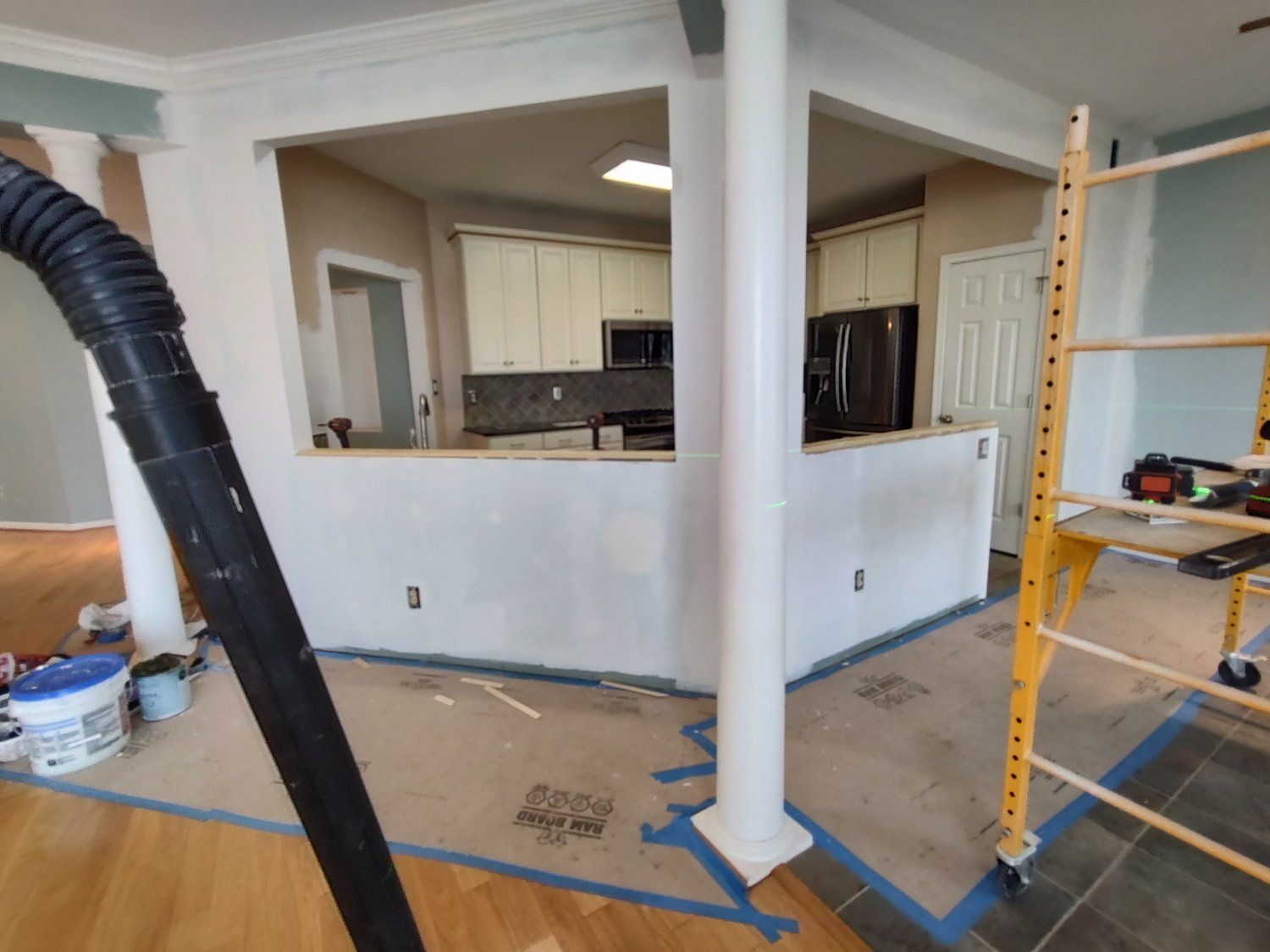 Renovation in progress: White walls, kitchen visible, scaffolding, black vacuum hose, and drop cloths on floor.